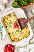 A baked egg casserole with spinach and cherry tomatoes in a white dish, partially served with a wooden spatula. Fresh spinach and tomatoes are nearby.