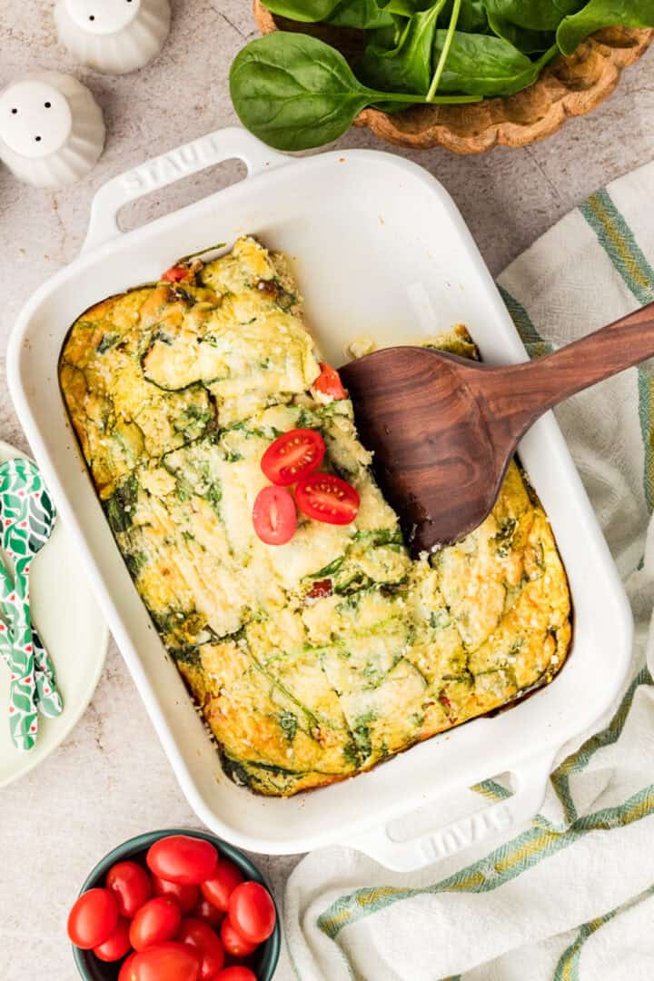 A baked egg casserole with spinach and cherry tomatoes in a white dish, partially served with a wooden spatula. Fresh spinach and tomatoes are nearby.