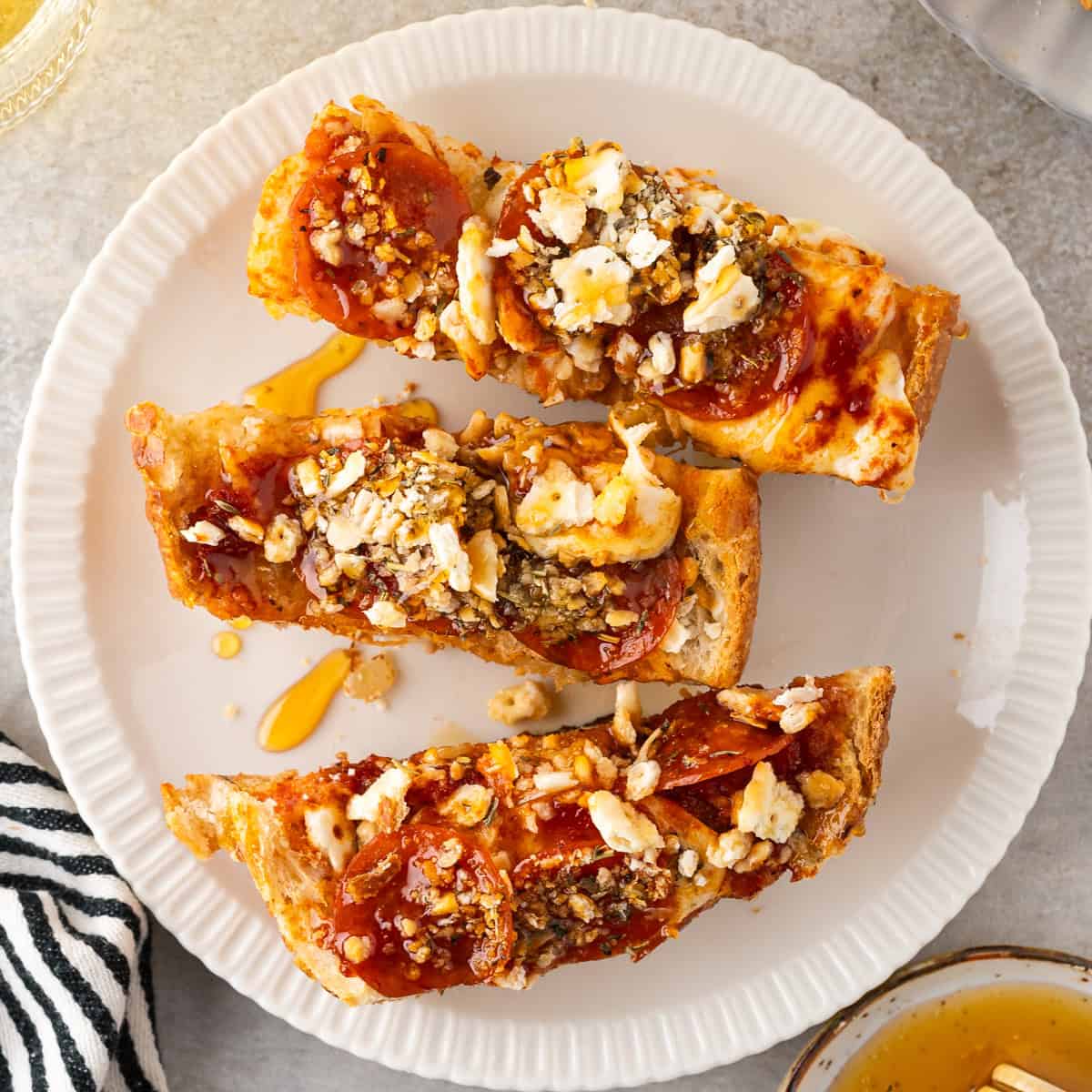 Three slices of toasted French Bread Pizza topped with pepperoni, crumbled cheese, and a drizzle of honey are arranged on a white plate.