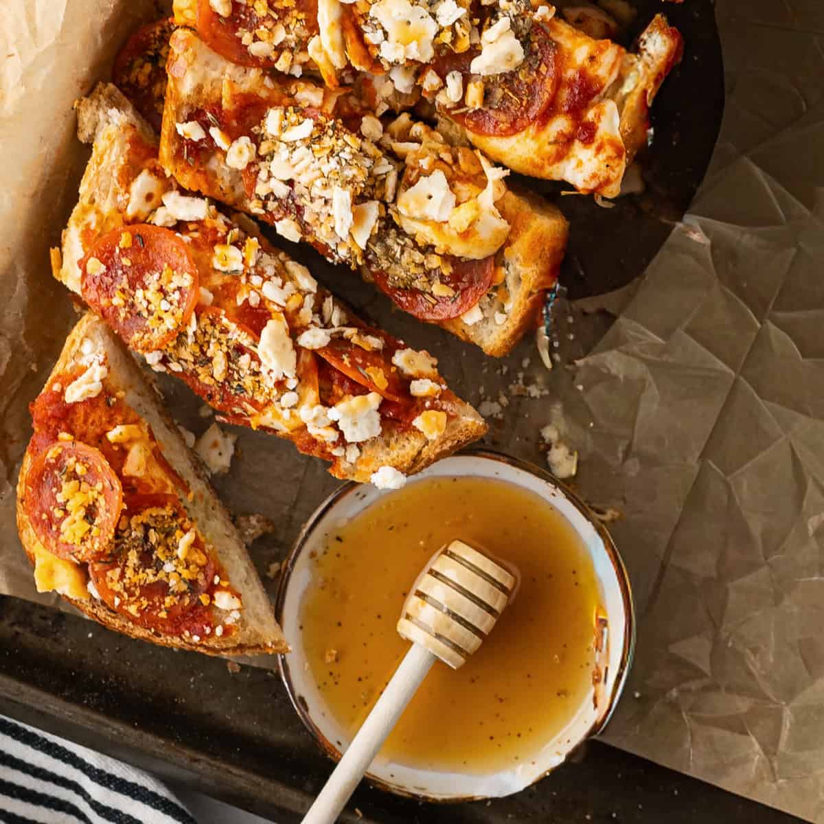 Slices of focaccia bread topped with pepperoni, cheese, and herbs are arranged on parchment paper next to a small bowl of honey with a honey dipper.