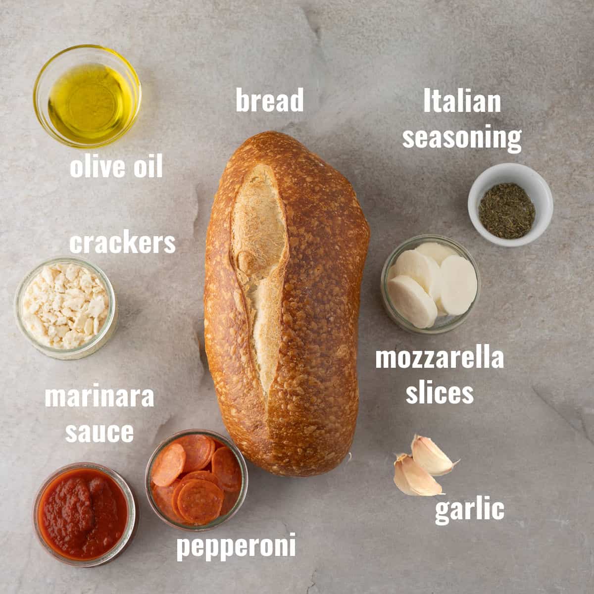 A loaf of bread surrounded by bowls of olive oil, crackers, Italian seasoning, mozzarella slices, marinara sauce, pepperoni, and garlic on a gray surface.
