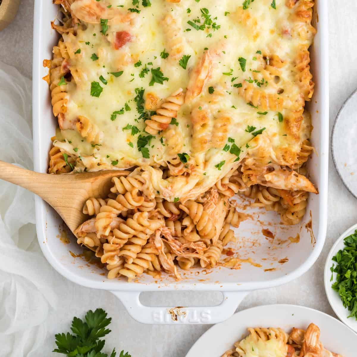 A baked pasta casserole with rotini, cheese, tomato sauce, and herbs in a white dish, with a wooden spoon serving a portion.