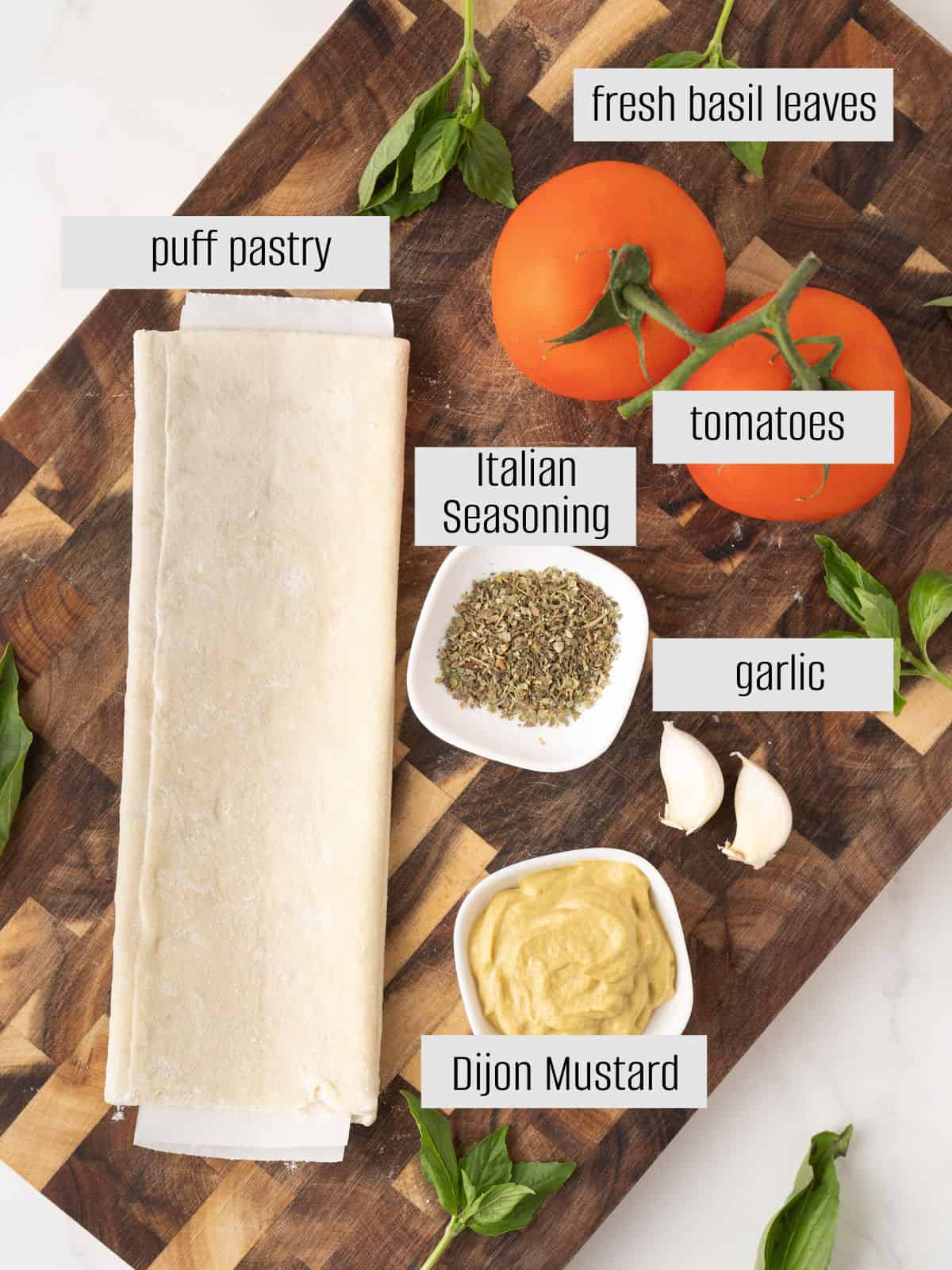 A wooden board with puff pastry, fresh basil leaves, tomatoes, Italian seasoning, garlic cloves, and Dijon mustard arranged in small piles or bowls.