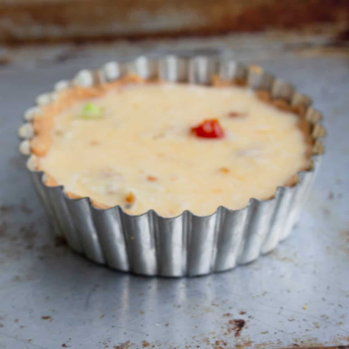 A small quiche with a golden crust and visible vegetable pieces in a fluted metal baking tin, placed on a worn metal surface.
