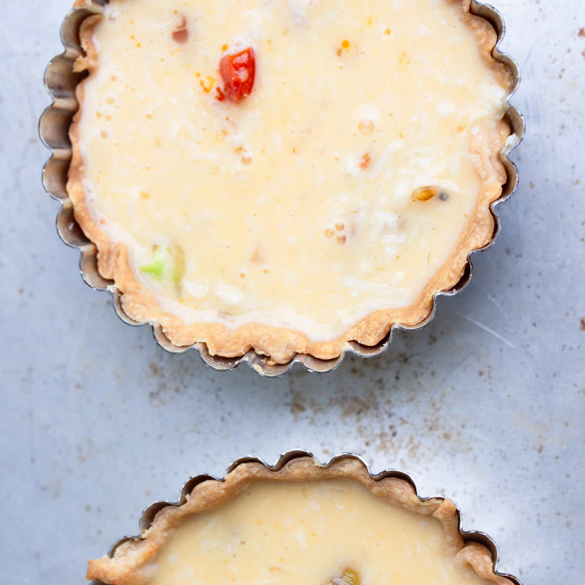 Two unbaked mini quiches in fluted metal tart pans on a light-colored surface; the filling appears creamy with visible vegetables.