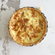 A baked quiche with a golden brown crust and cheesy filling sits in a round pan on a textured, white surface, ready to serve or perfect for those learning how to freeze quiche for later enjoyment.
