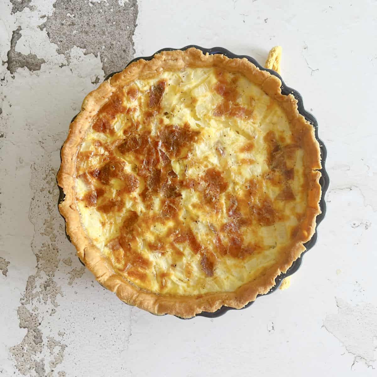 A baked quiche with a golden brown crust and cheesy filling sits in a round pan on a textured, white surface, ready to serve or perfect for those learning how to freeze quiche for later enjoyment.