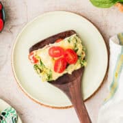 A slice of vegetable casserole topped with cherry tomato halves on a wooden spatula over a light green plate.
