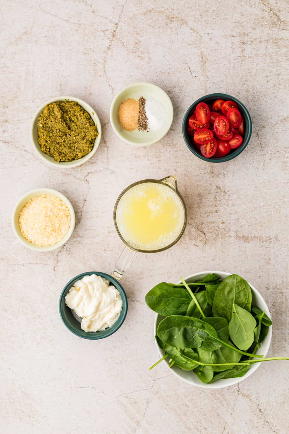 Six small bowls containing pesto, grated cheese, mayonnaise, baby spinach, cherry tomatoes, and seasonings, arranged on a light marble surface with a measuring cup of liquid egg whites in the center.