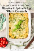 A baked ricotta and spinach egg white casserole in a white dish, garnished with basil, next to a fork, knife, and bowl of grape tomatoes.