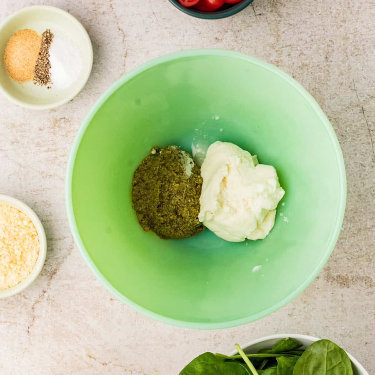 A green bowl containing pesto and ricotta, surrounded by small bowls of grated cheese, spices, cherry tomatoes, and fresh spinach on a light countertop.