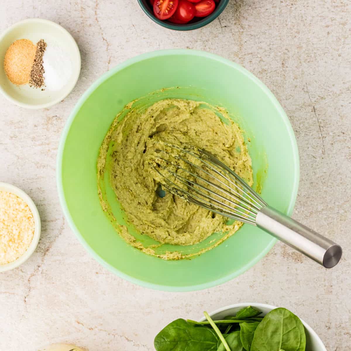 A green mixing bowl with a metal whisk contains a creamy green mixture, surrounded by bowls of seasonings, grated cheese, cherry tomatoes, and fresh spinach.