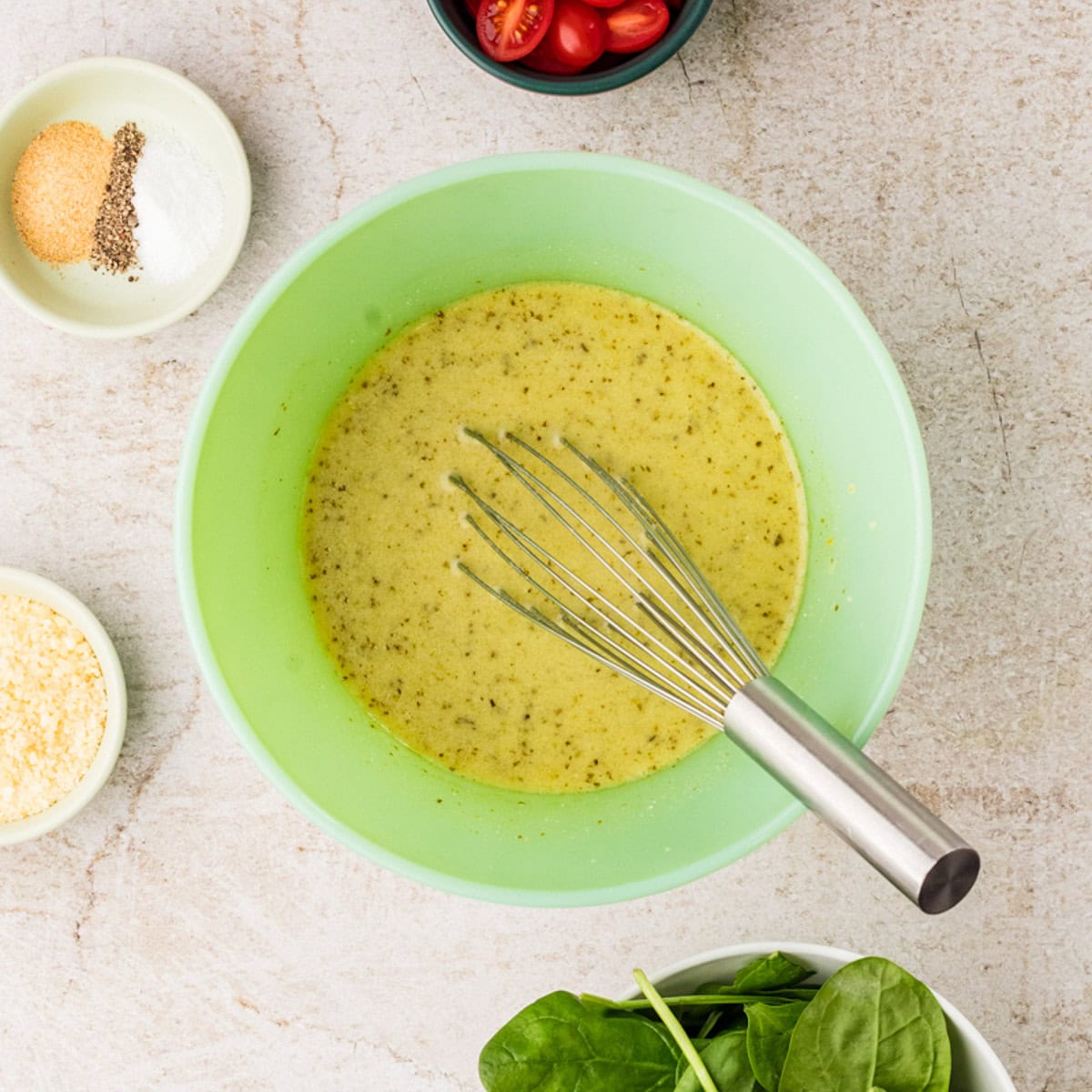 A green mixing bowl with a whisk in yellow-green dressing, perfect for preparing an egg white casserole, surrounded by bowls of seasoning, grated cheese, cherry tomatoes, and fresh spinach.