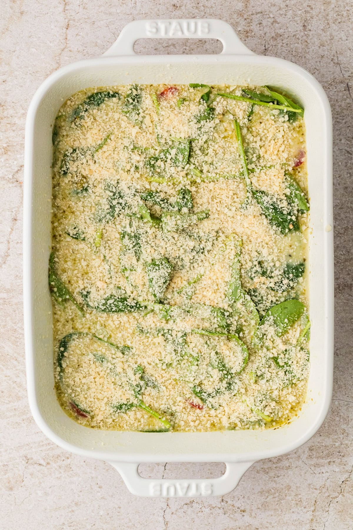 A white baking dish filled with a creamy egg white casserole mixture of spinach and topped with breadcrumbs, ready to be baked.
