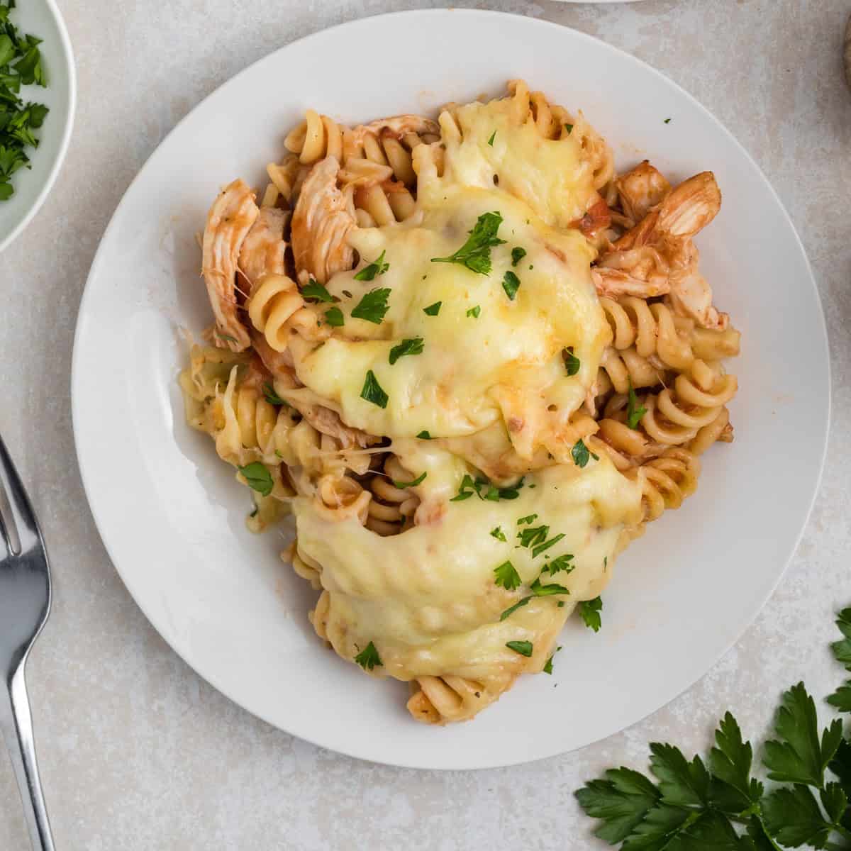 A white plate of rotini pasta with shredded chicken, topped with melted cheese and chopped parsley, sits on a light surface next to a fork and fresh parsley.