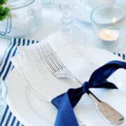 A neatly folded white napkin with a fork tied by a navy blue ribbon rests on a white plate, capturing the essence of an evening ritual, with candles and glassware softly illuminating the background.