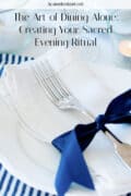 Close-up of a place setting with a white napkin, silver fork, and dark blue ribbon on a white plate; text reads The Art of Dining Alone: Creating Your Sacred Evening Ritual.