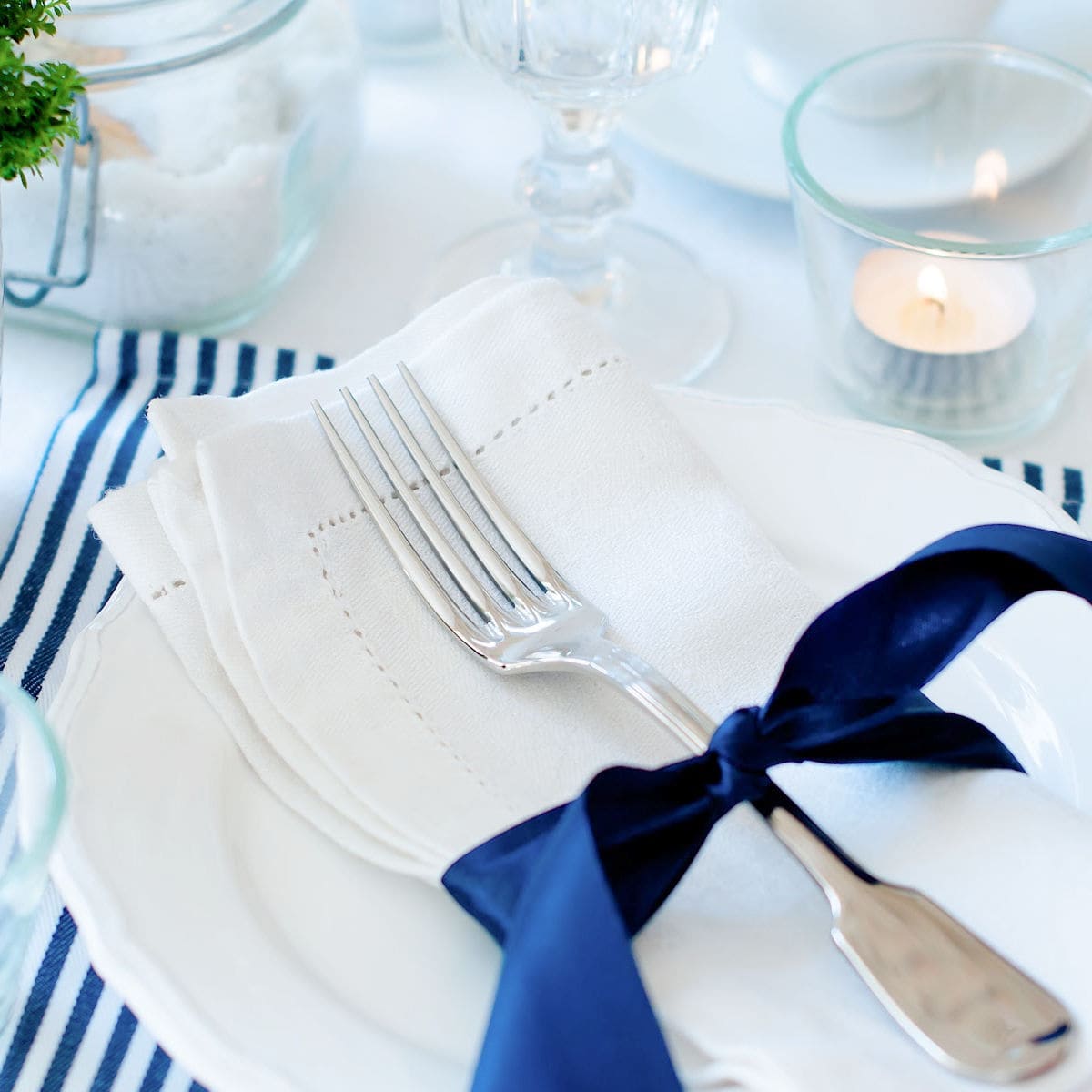 A neatly folded white napkin with a fork tied by a navy blue ribbon rests on a white plate, capturing the essence of an evening ritual, with candles and glassware softly illuminating the background.