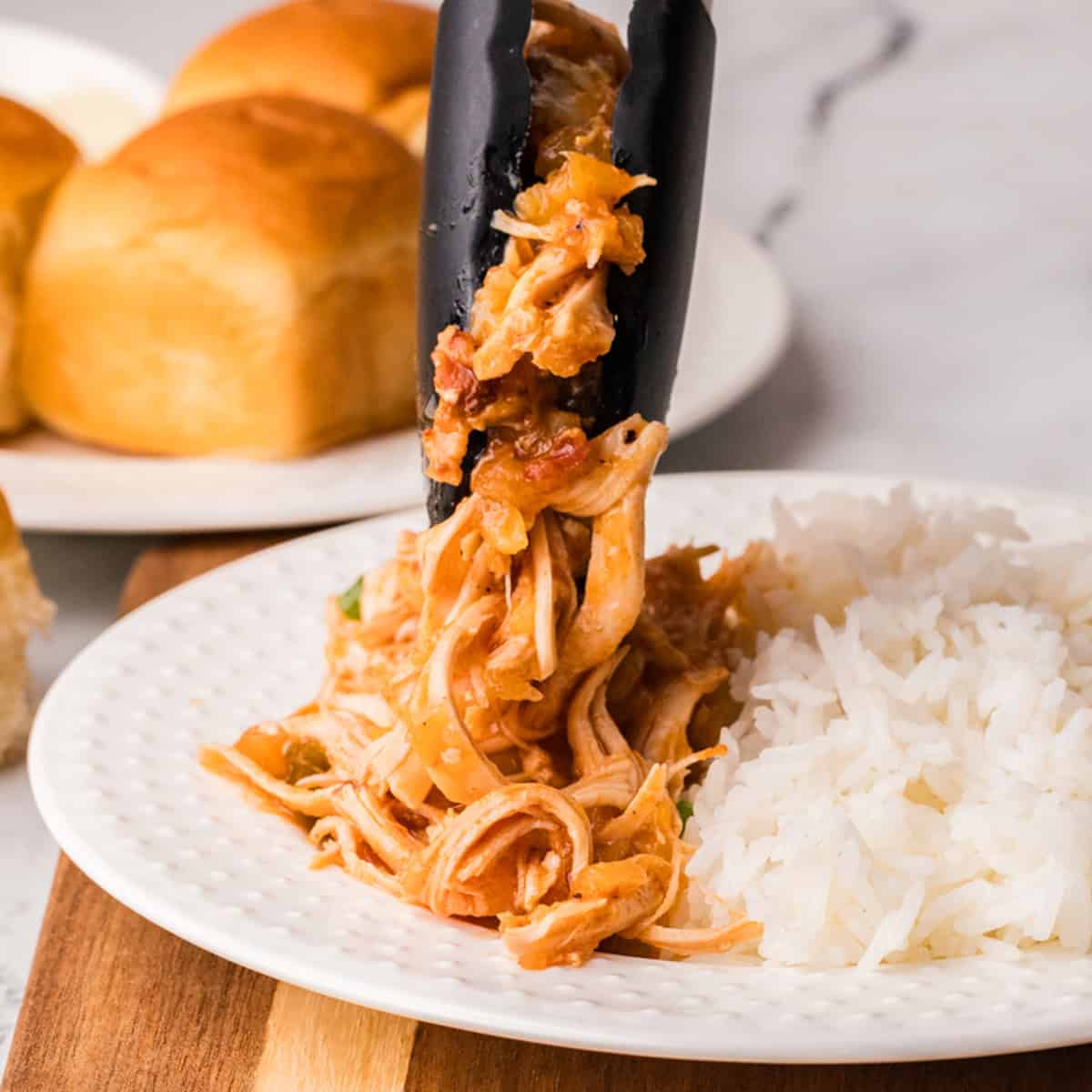 A serving of shredded chicken with sauce is being placed next to a portion of white rice on a plate; dinner rolls are visible in the background.