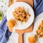 White plate with shredded chicken in sauce over rice, topped with chopped green onions, served with a bread roll on a wooden board, blue cloth nearby.
