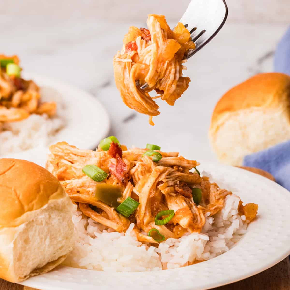 A plate with shredded chicken in sauce over white rice, garnished with green onions, next to a dinner roll; a fork holds up a bite of chicken.