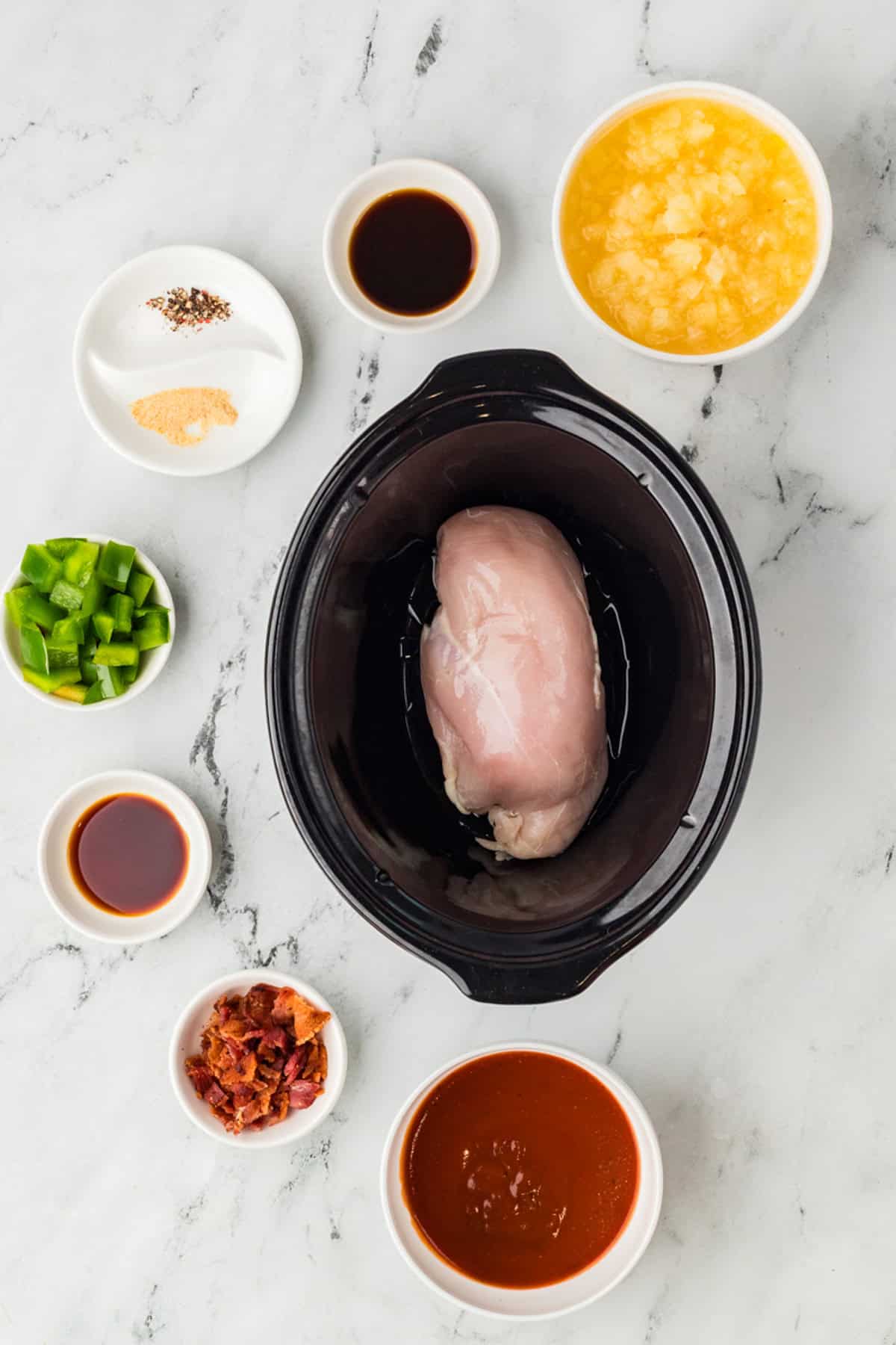 A raw chicken breast in a slow cooker surrounded by small bowls of pineapple, barbecue sauce, bacon, green peppers, seasonings, and sauces on a marble surface.