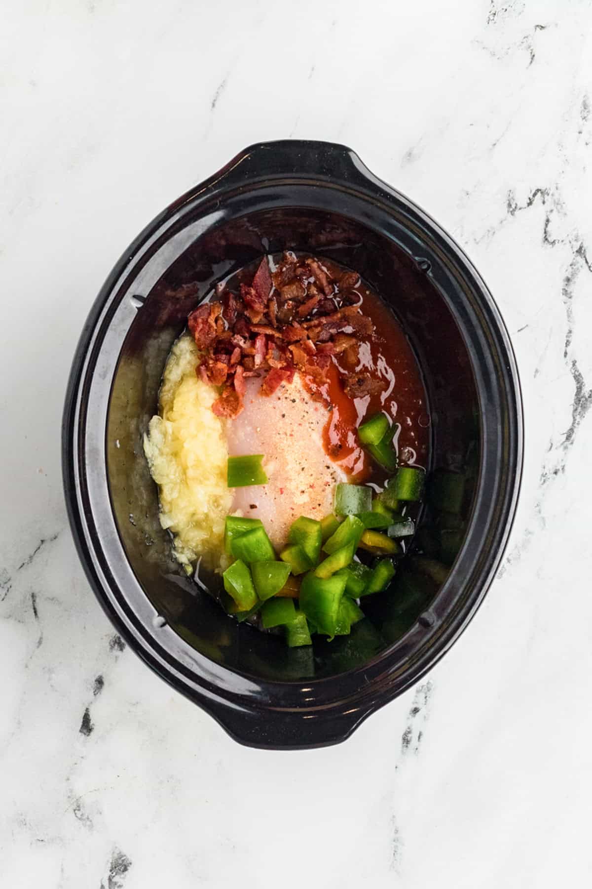 A slow cooker containing shredded potatoes, chopped green bell pepper, cooked bacon, shredded cheese, and a creamy sauce, ready to be mixed on a marble countertop.
