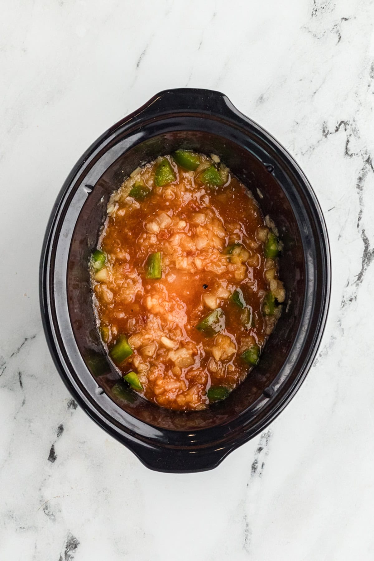 A black slow cooker containing a mixture of chopped green peppers, onions, and tomato sauce sits on a white marble surface.