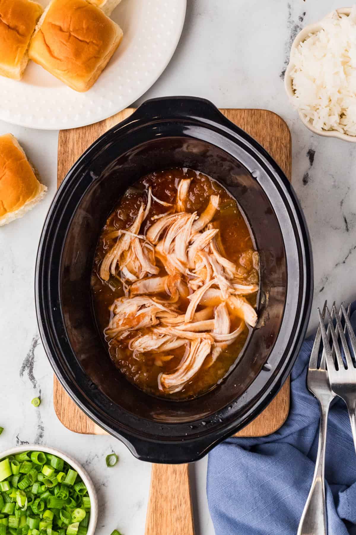 Shredded chicken in sauce inside a slow cooker, surrounded by green onions, rice, dinner rolls, and forks on a marble surface.