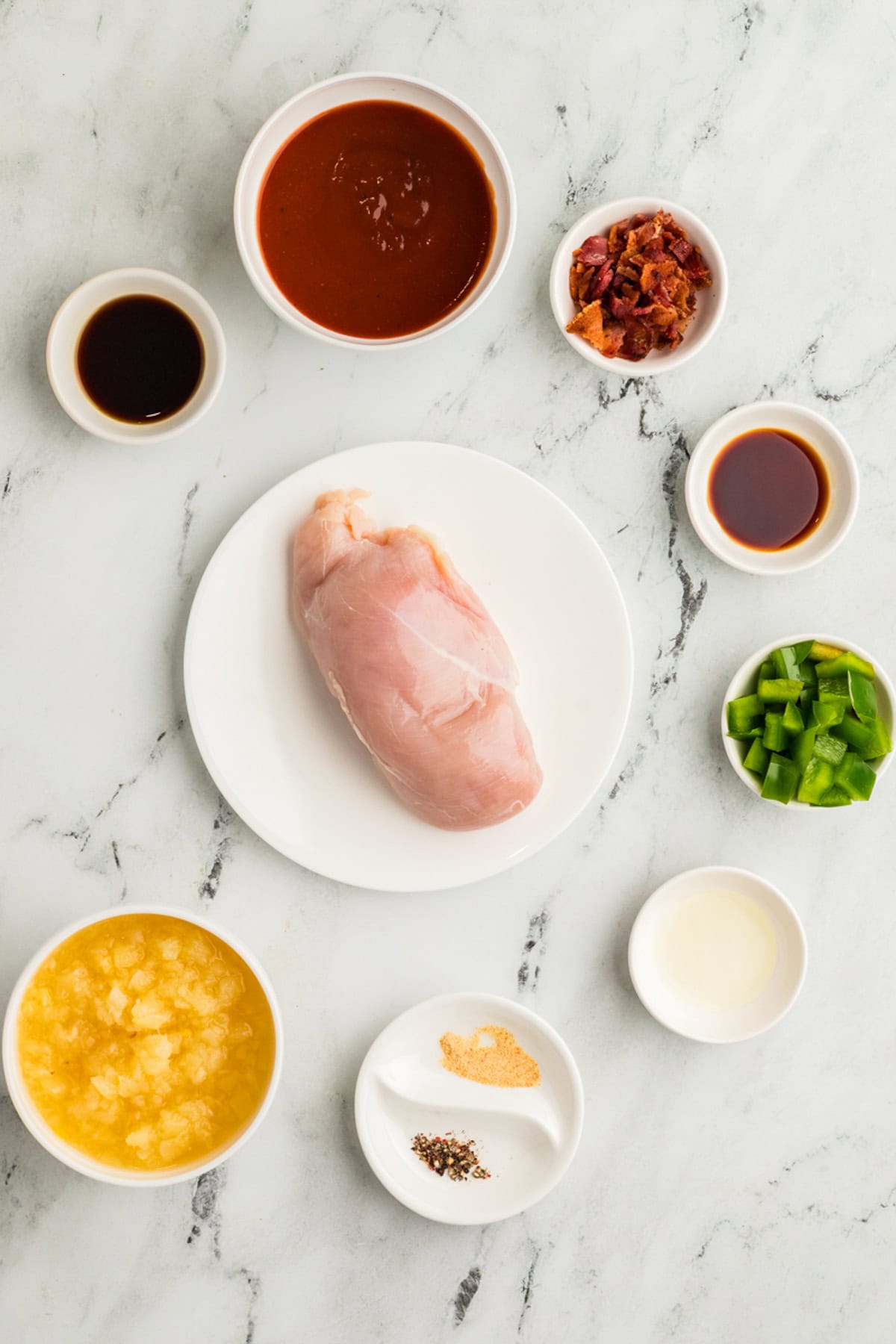 A raw chicken breast on a plate surrounded by small bowls containing barbecue sauce, chopped bacon, soy sauce, pineapple, green bell pepper, vinegar, spices, and Worcestershire sauce.