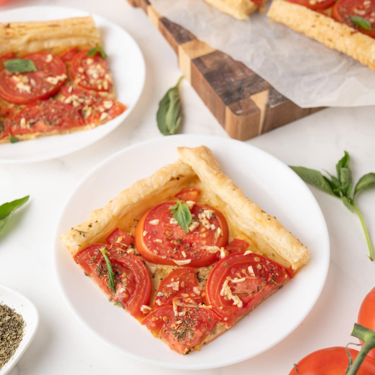 A slice of tomato tart with puff pastry, herbs, and fresh basil on a white plate, surrounded by ingredients and another serving in the background.