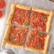 A rectangular tomato tart with puff pastry, sliced tomatoes, herbs, and garlic is cut into four pieces and placed on parchment paper.