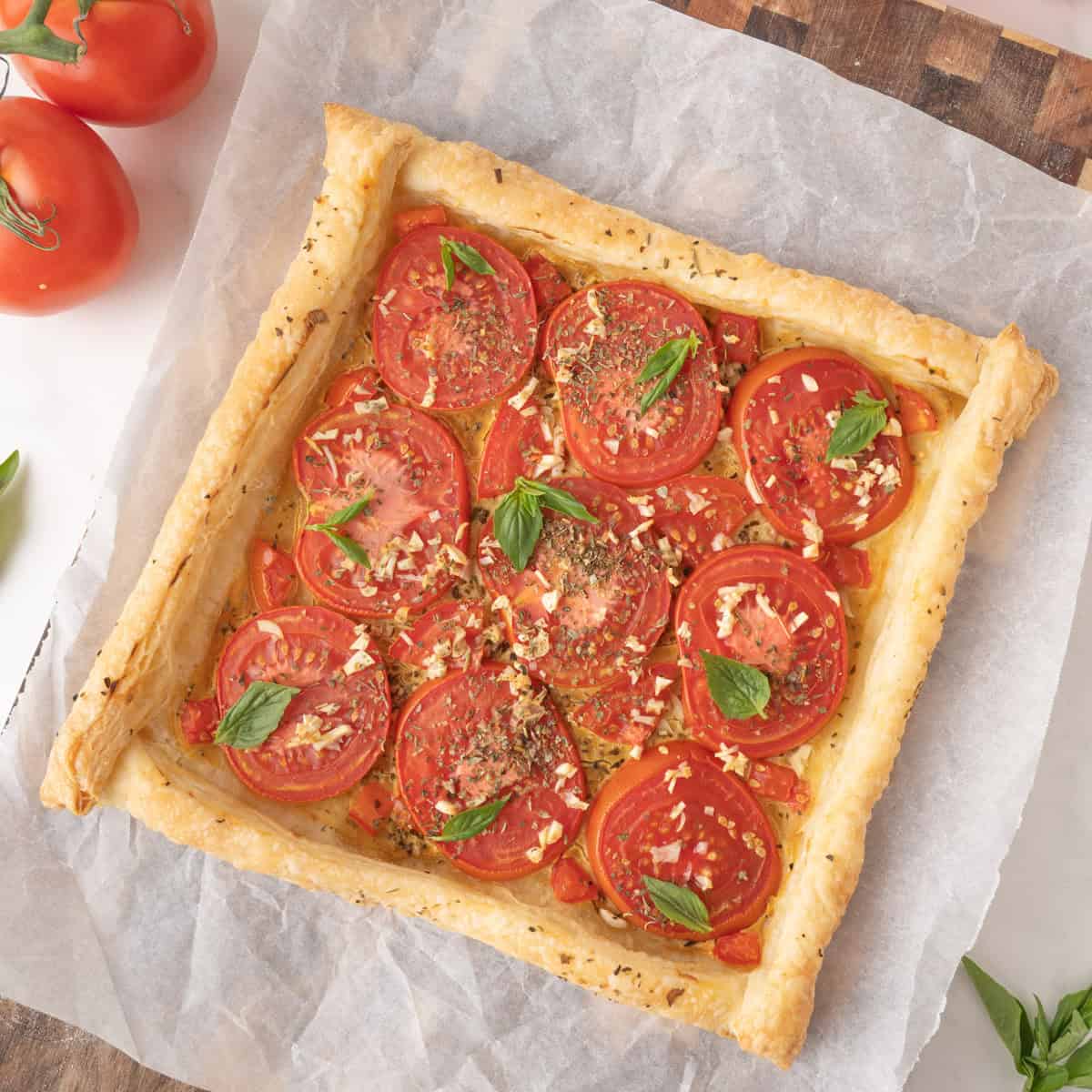 A square puff pastry topped with sliced tomatoes, chopped garlic, herbs, and basil leaves, placed on parchment paper on a wooden board.