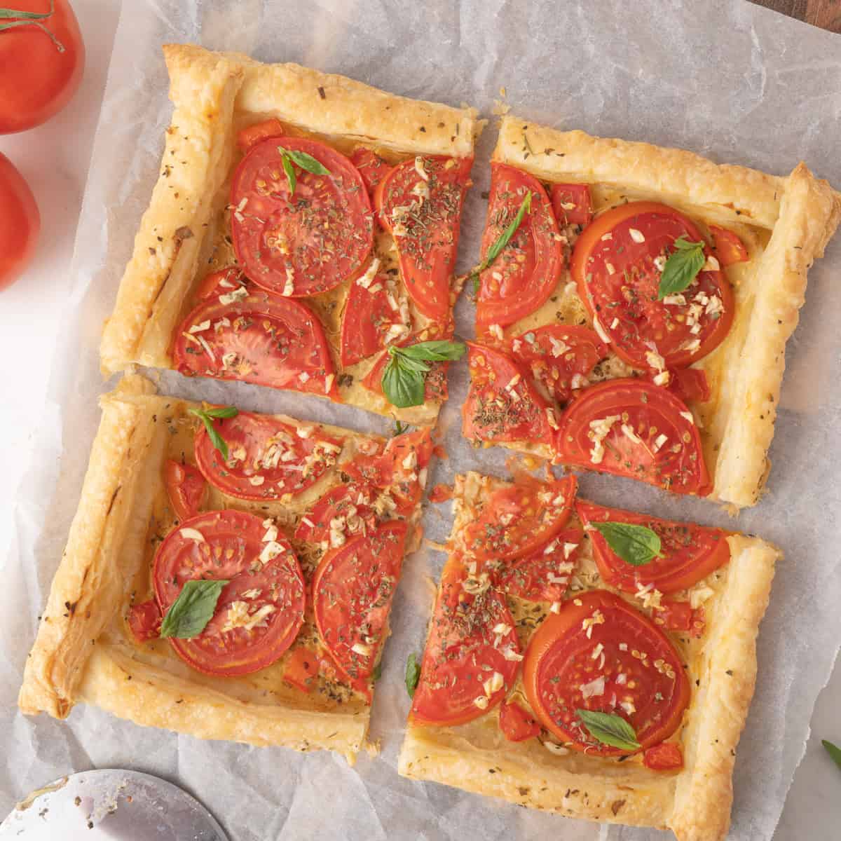 A rectangular tomato tart with puff pastry, sliced tomatoes, herbs, and garlic is cut into four pieces and placed on parchment paper.