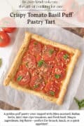 A square puff pastry tart topped with sliced tomatoes and fresh basil sits on parchment paper, surrounded by whole tomatoes and herbs.