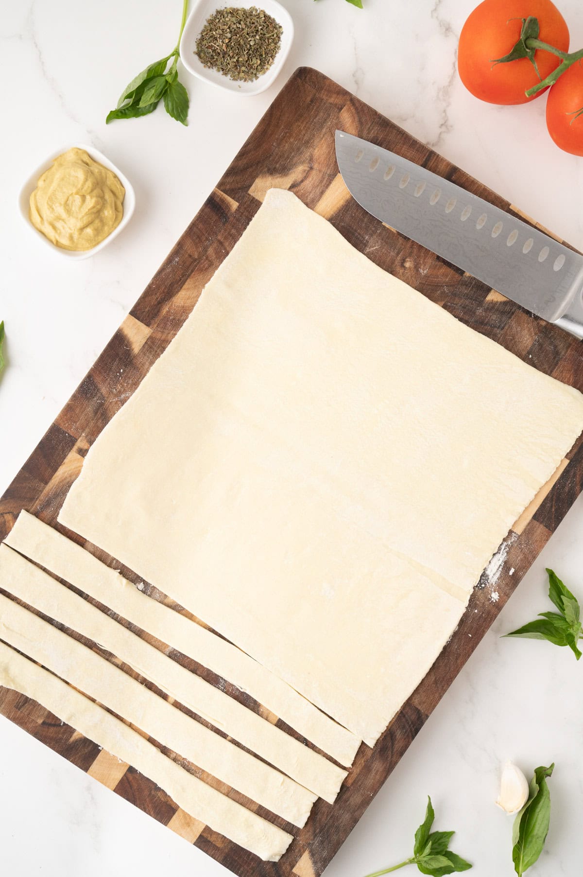A sheet of dough is partially cut into strips on a wooden cutting board, surrounded by a knife, mustard, herbs, garlic, and tomatoes.