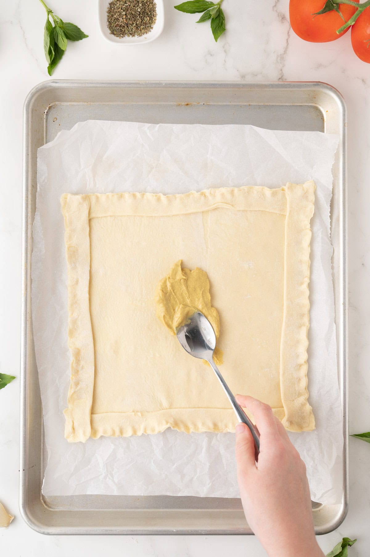A hand spreads mustard onto a square sheet of pastry dough on a parchment-lined baking tray, surrounded by fresh basil, tomatoes, and a bowl of dried herbs.