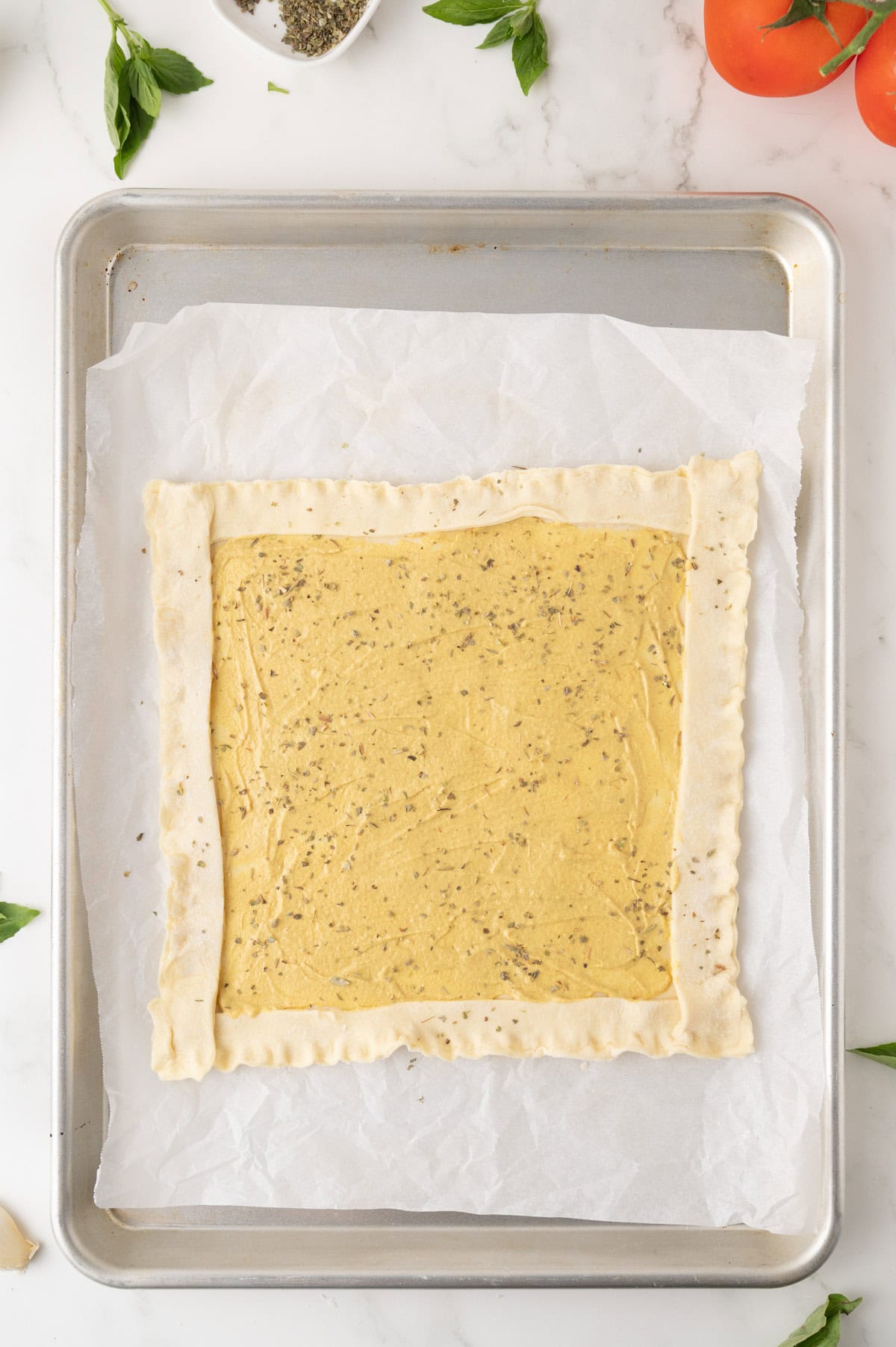 A square sheet of dough with folded edges and an herbed filling sits on parchment paper on a baking tray, surrounded by fresh basil and tomatoes.