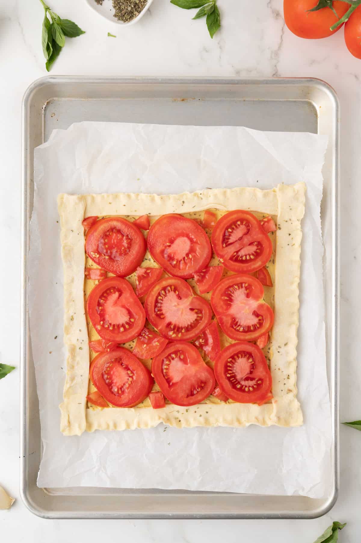 Unbaked tomato tart on parchment-lined baking sheet, topped with fresh tomato slices and herbs, with edges of pastry folded inward.