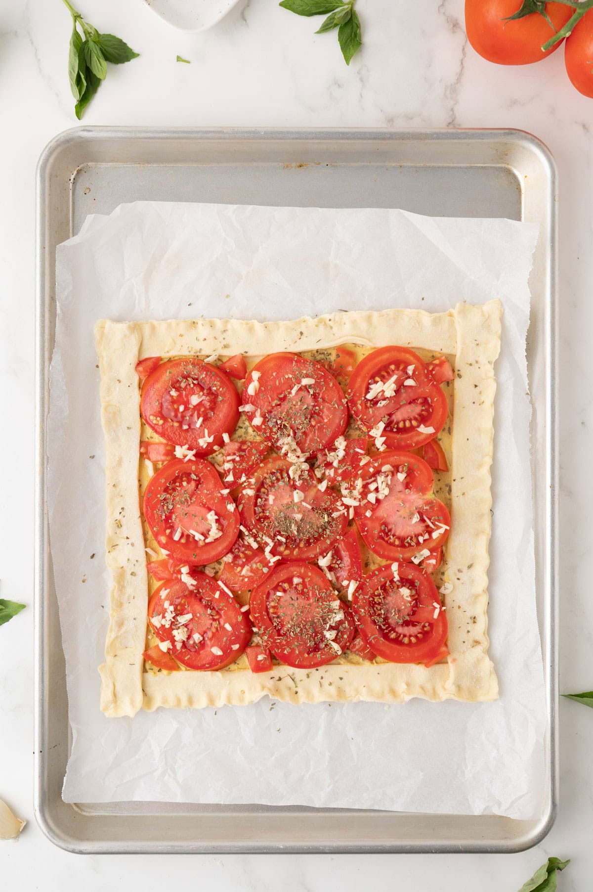 A square tomato tart with herbs and cheese on puff pastry, placed on parchment paper atop a baking sheet.