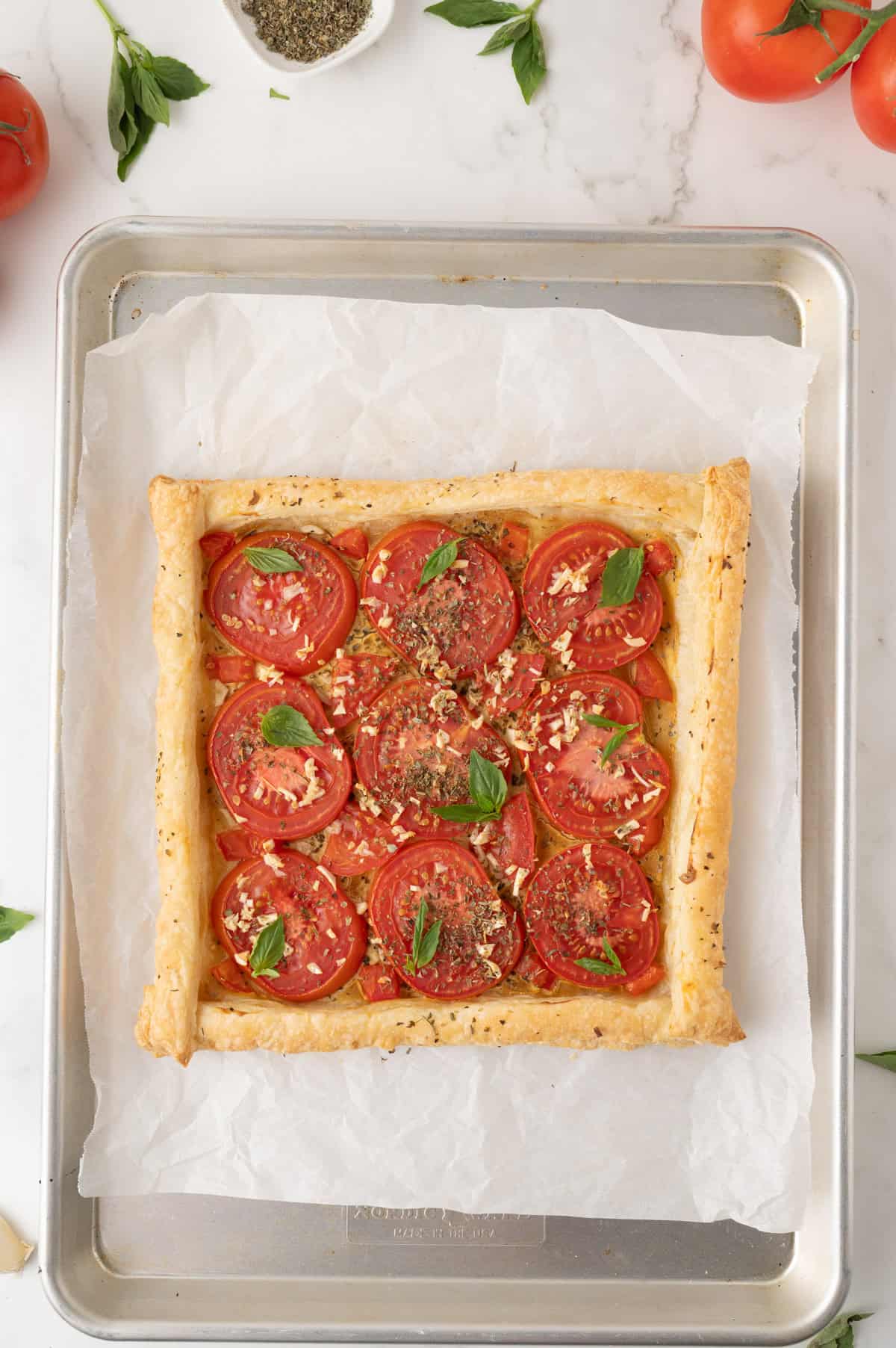 A rectangular tomato tart with puff pastry, sliced tomatoes, herbs, and basil leaves on parchment paper atop a baking tray.