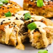 Three golden brown crab cakes topped with creamy sauce, black sesame seeds, and chopped green onions are shown on a plate.