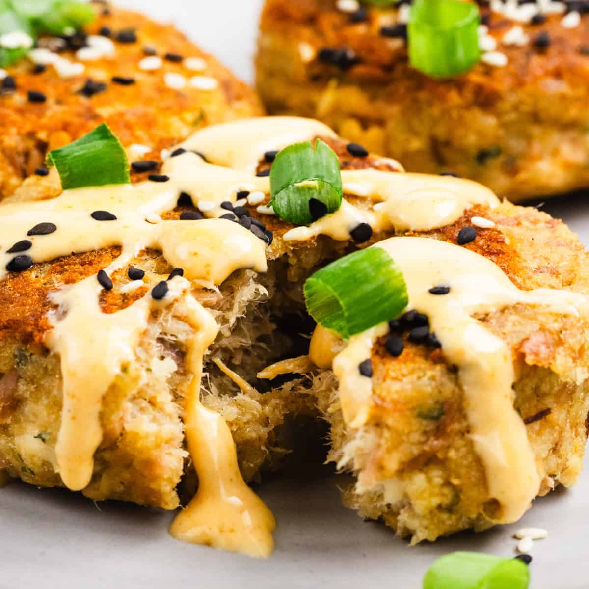 Three golden brown crab cakes topped with creamy sauce, black sesame seeds, and chopped green onions are shown on a plate.