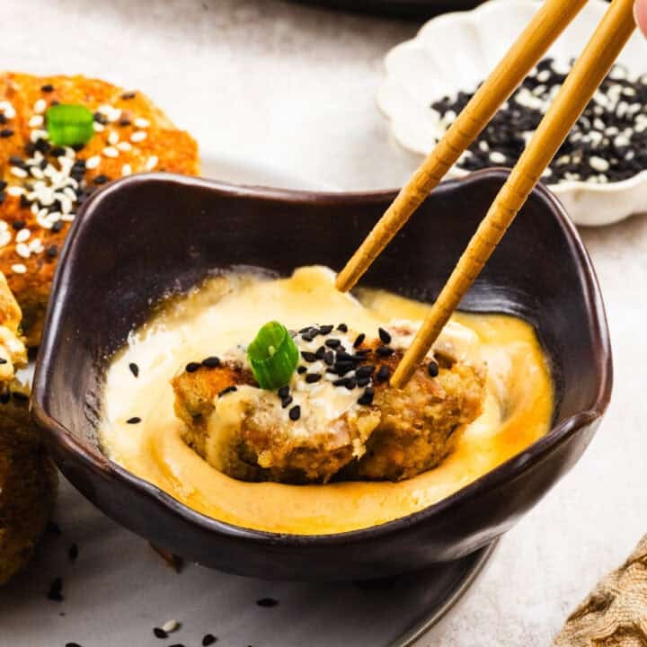 A pair of chopsticks holds a pan fried tuna cake dipped in a spicy mayo, garnished with black and white sesame seeds and a green onion slice in a black bowl.