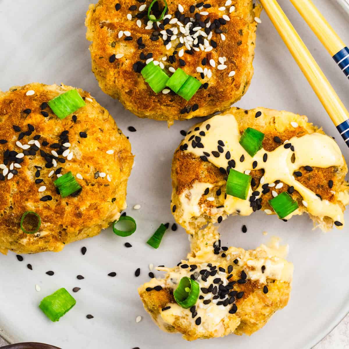Three golden-brown tuna patties topped with sesame seeds and chopped green onions are on a plate, with one patty broken open to show a creamy filling; chopsticks rest nearby.