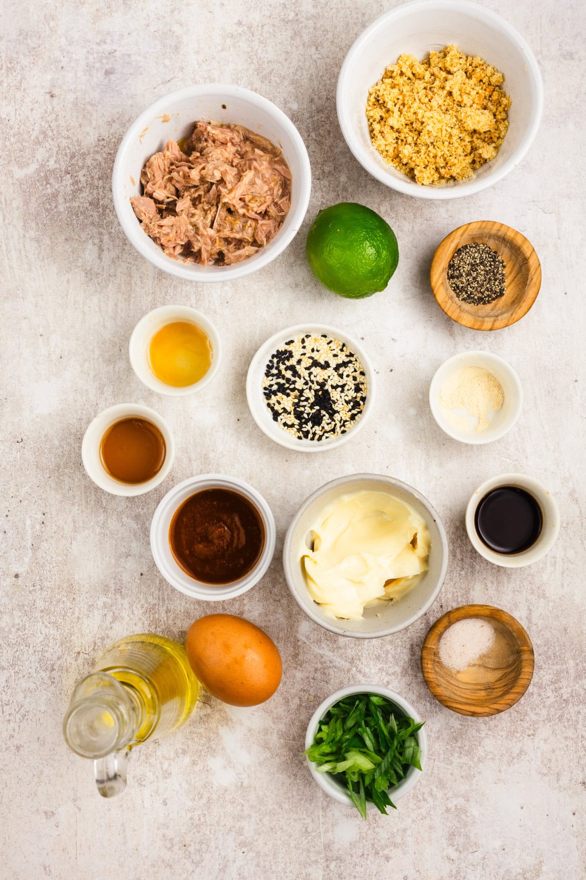 A variety of labeled ingredients in small bowls, including tuna, mayonnaise, egg, green onions, spices, lime, oil, and seasonings, arranged on a light surface.