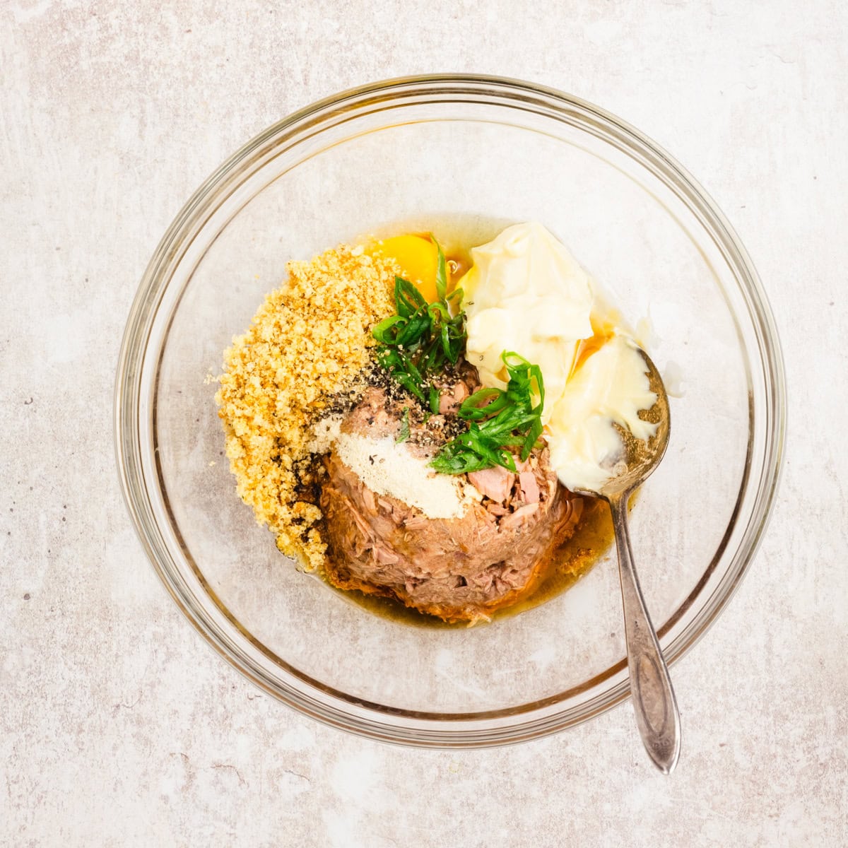 A glass bowl containing tuna, breadcrumbs, an egg, mayonnaise, chopped green onions, and seasoning, with a spoon on the side.