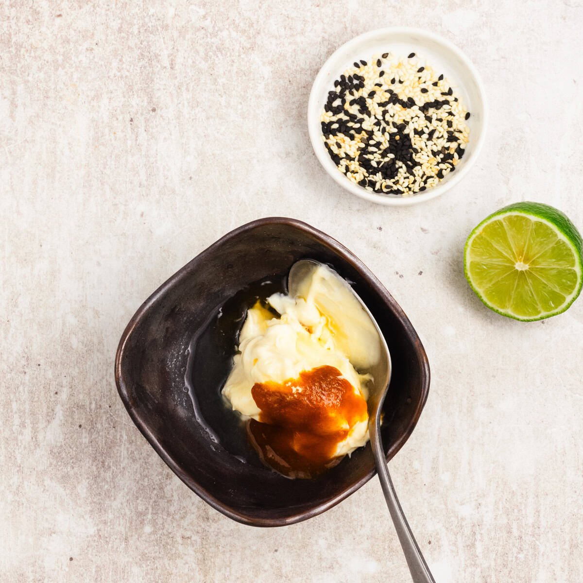 A bowl with mayonnaise and red sauce, a spoon, a dish of sesame seeds, and half a lime on a light surface.