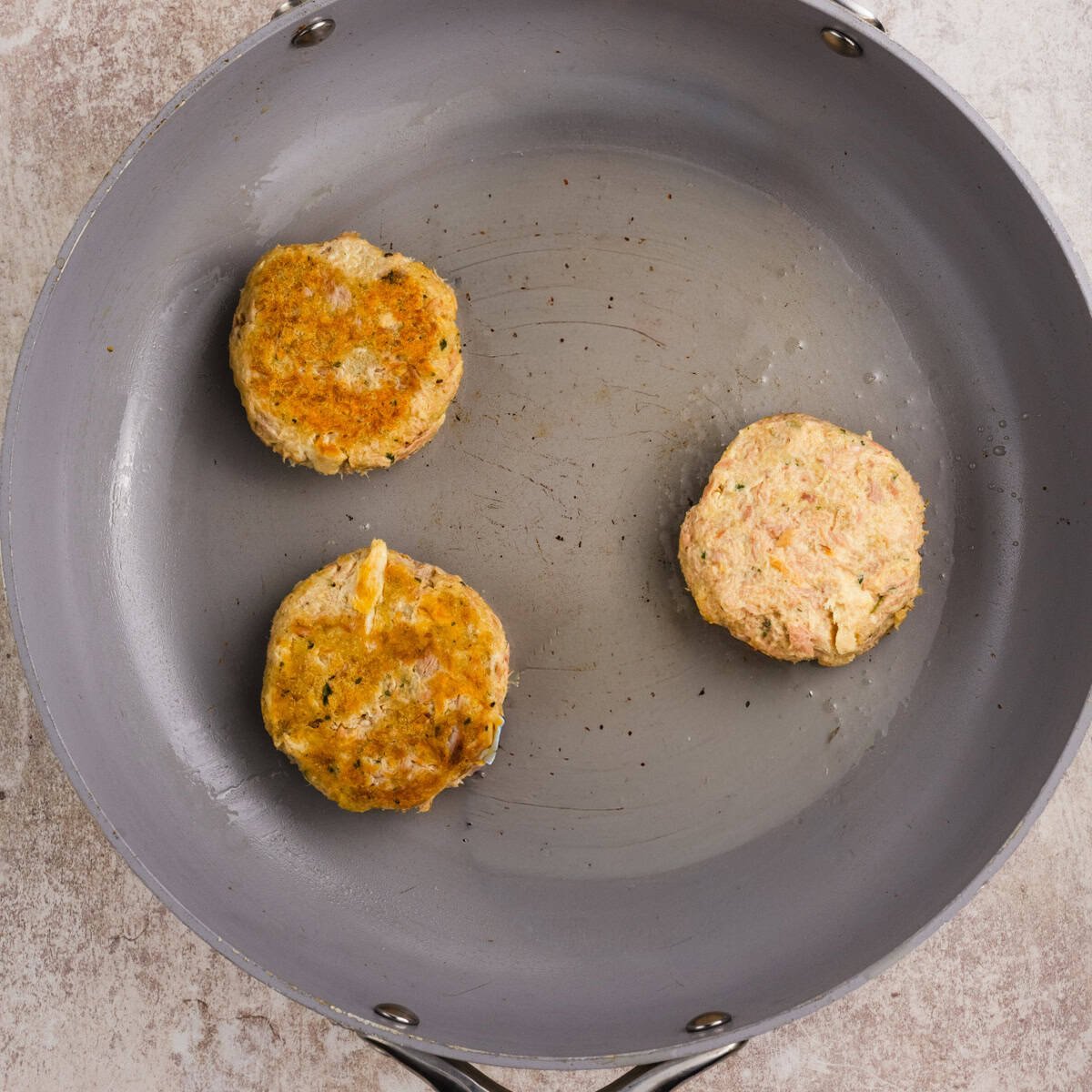 Three round patties are cooking in a gray frying pan; two are browned and one is still pale, indicating it hasnt been flipped yet.