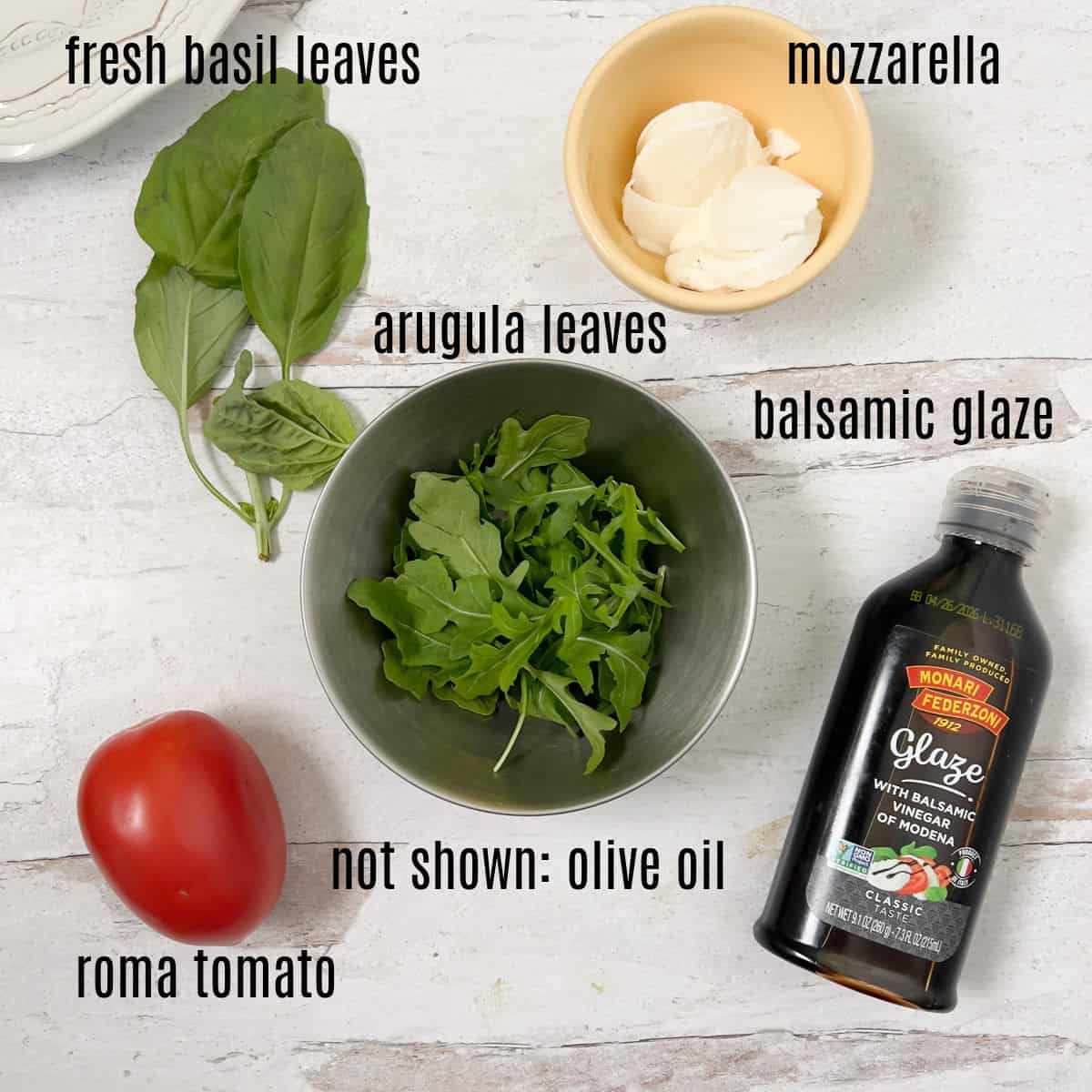 Ingredients for a salad are arranged on a white surface, including basil leaves, arugula, mozzarella, roma tomato, and balsamic glaze, with olive oil not shown.